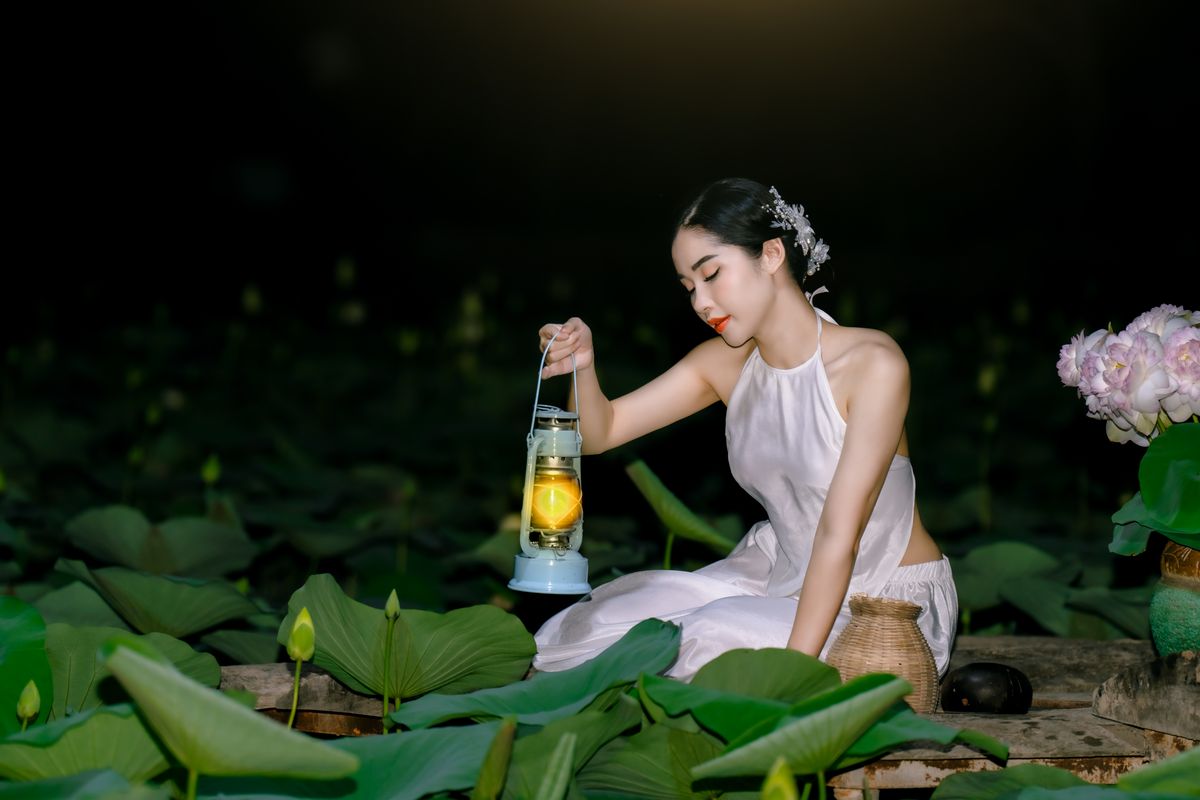 A young woman in traditional attire holds a lantern among lotus leaves at night.