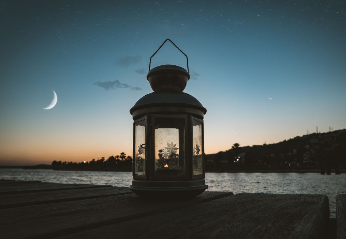Serene evening scene featuring a lantern on a dock by the water under a crescent moon and starry sky.