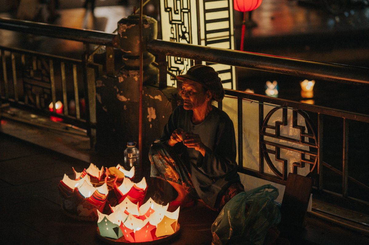 An elderly woman sits on the pavement selling paper lanterns under urban lights at night.