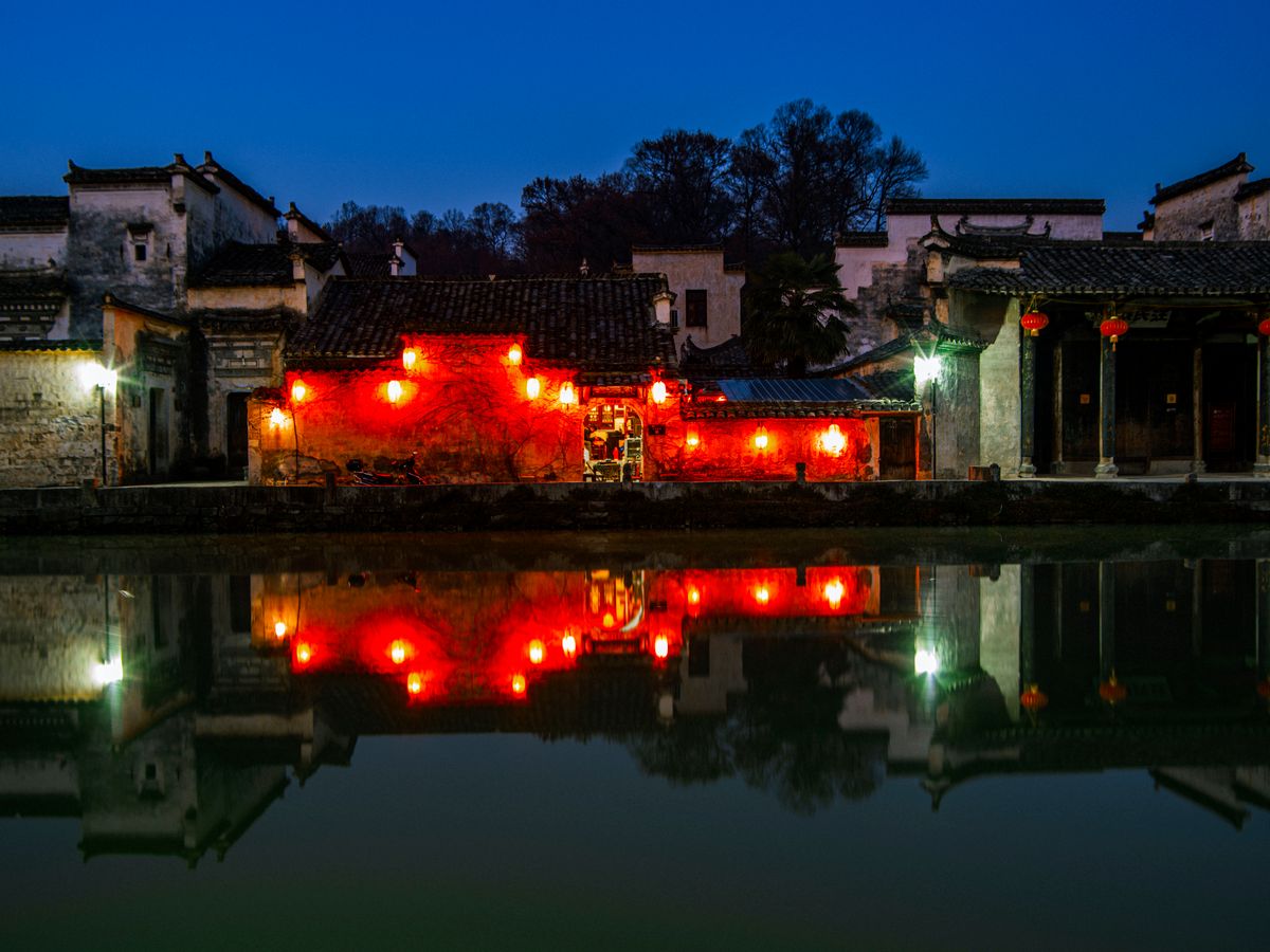 Illuminated Chinese architectural building reflecting on water during twilight with vibrant red lights.