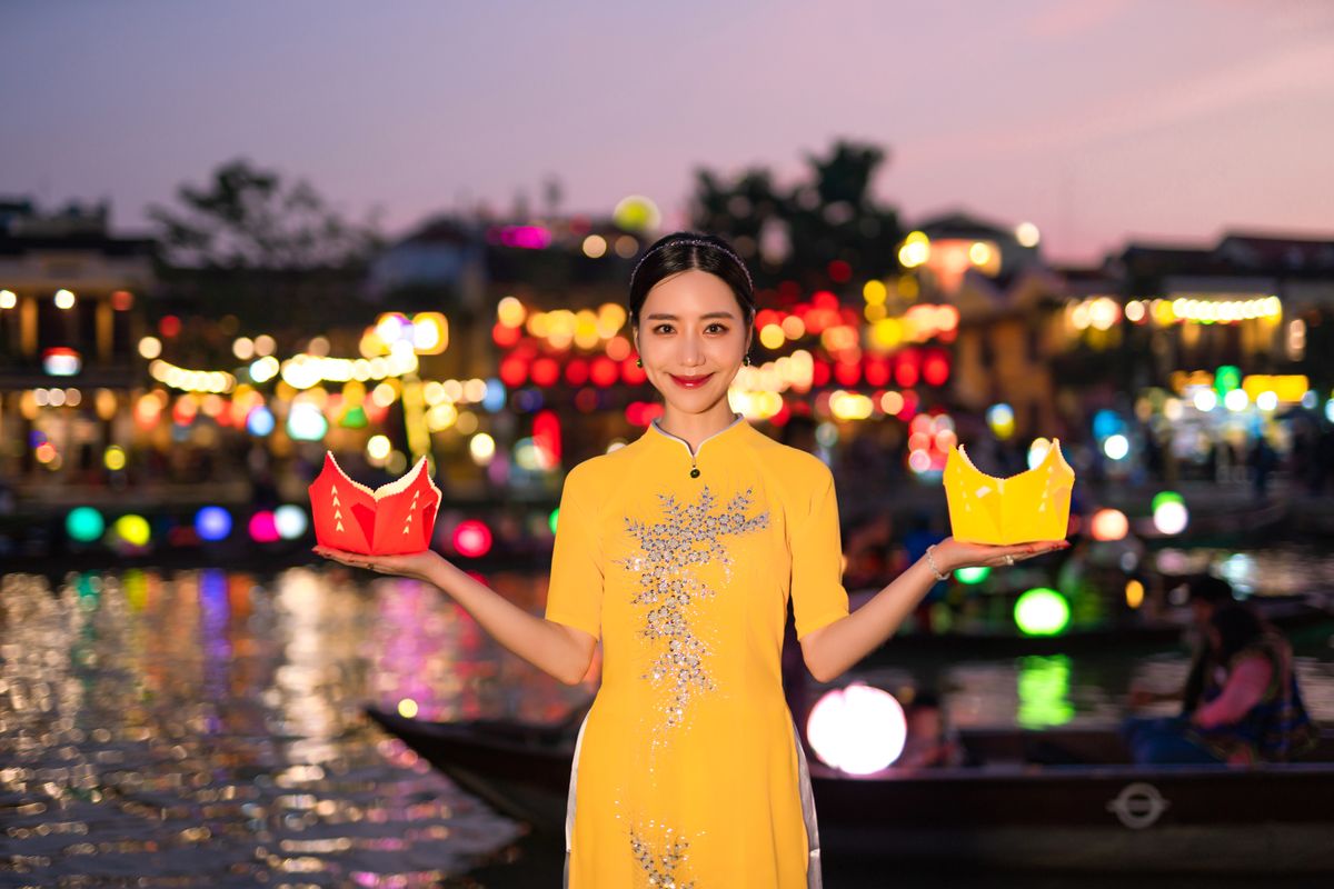 Woman in traditional dress holding lanterns at vibrant night festival by the water.