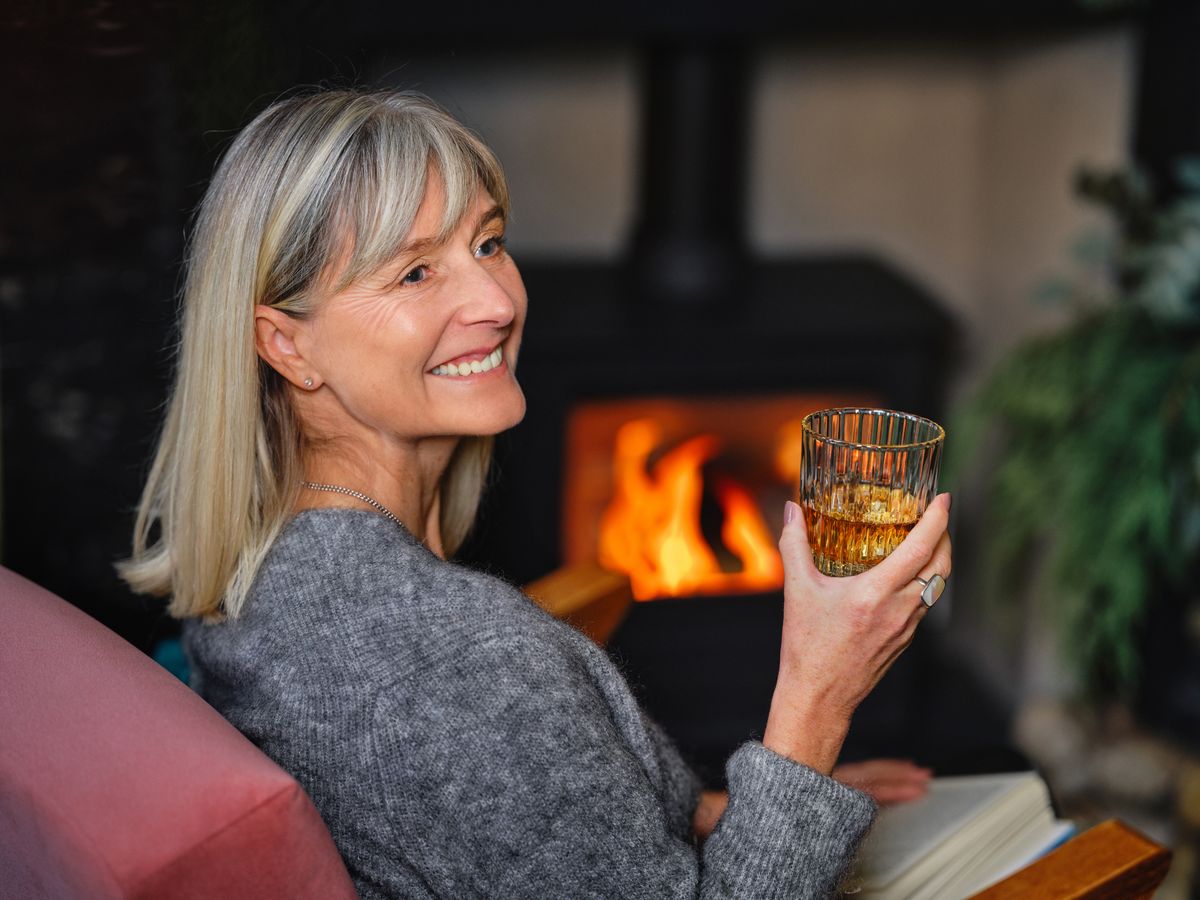 Senior woman enjoying a relaxing evening by the fireplace holding a glass of whiskey.