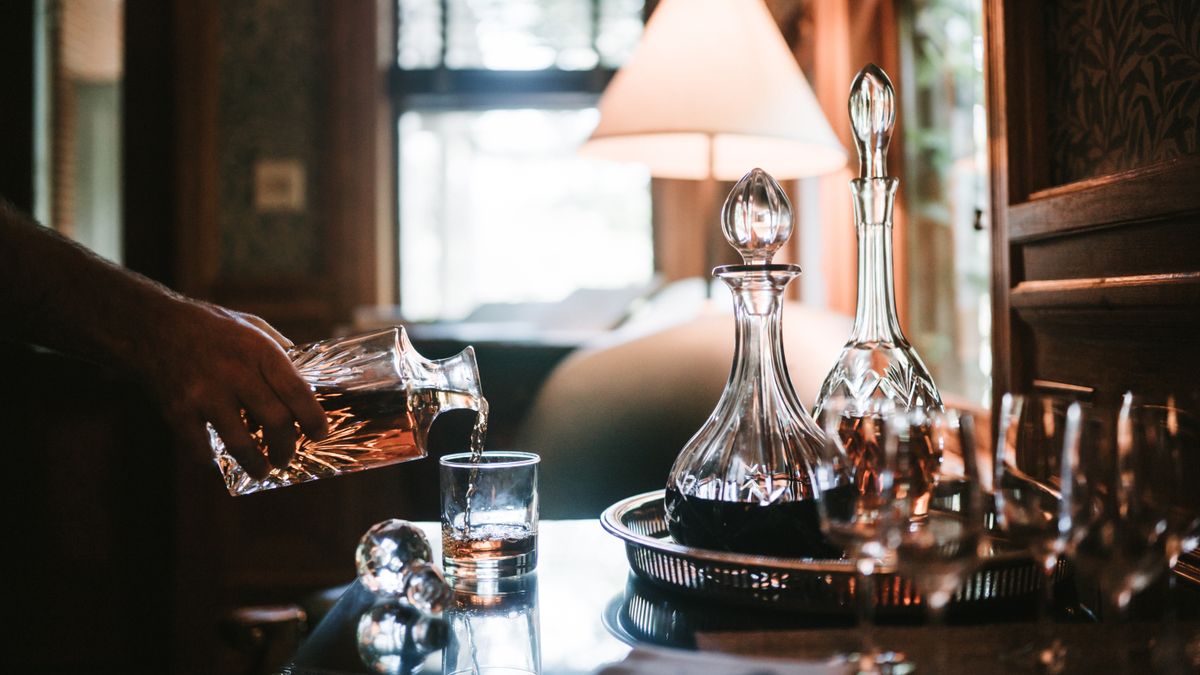 A hand pours whiskey from a crystal decanter into a glass in a cozy indoor setting.