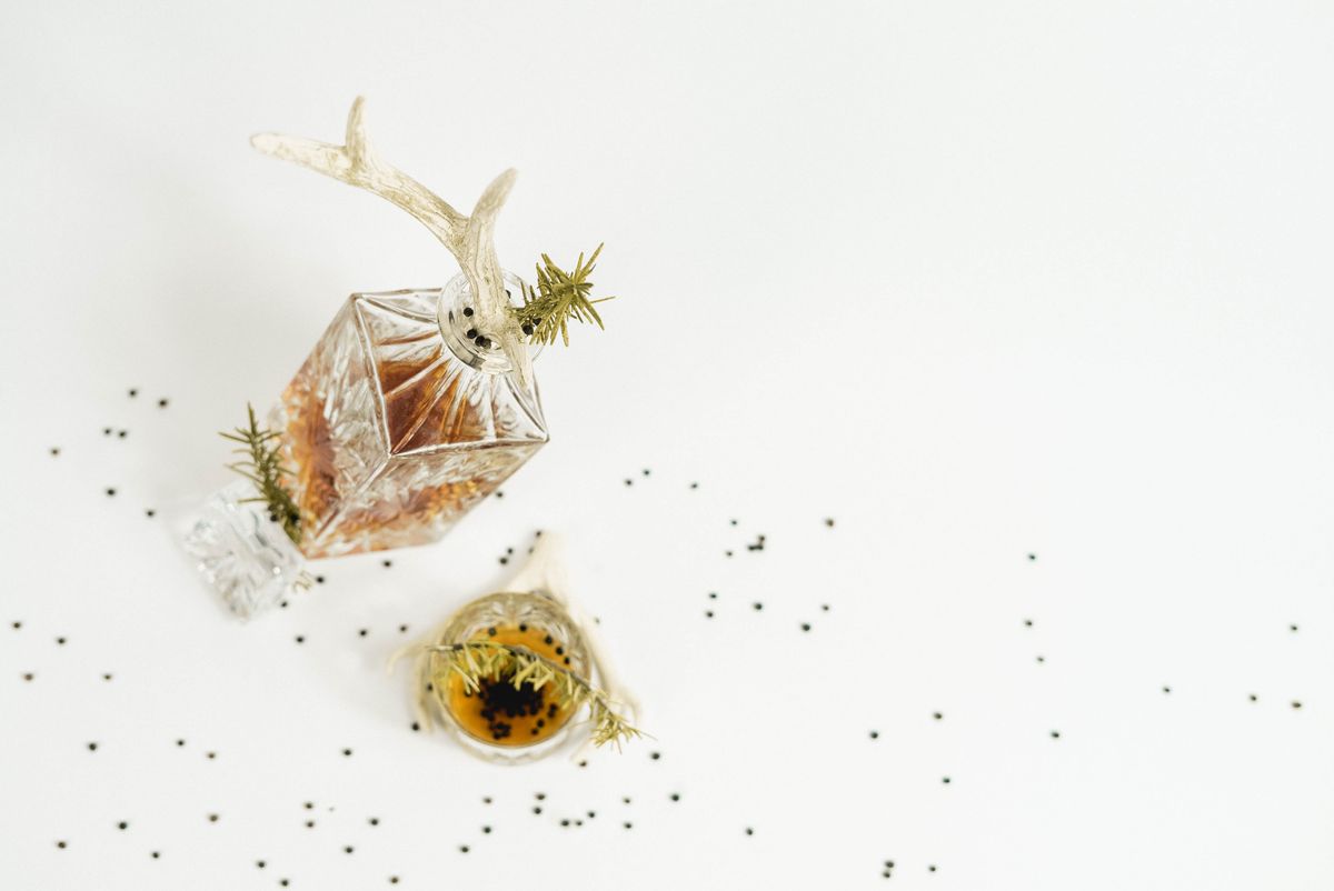 From above of carafe and glass filled with whiskey served with black peppercorns and green rosemary twigs on white background