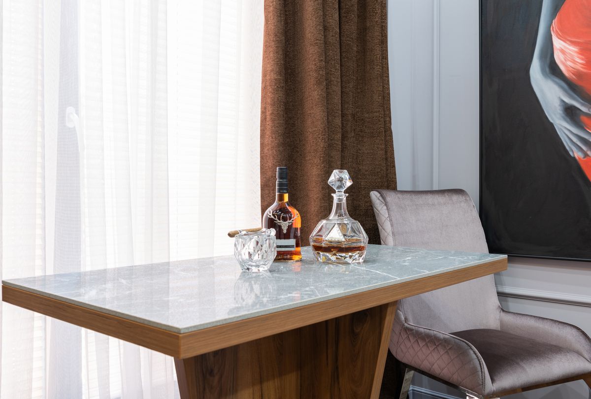 Bottle of liquor between decanter and glass with cigar on table against curtain in house