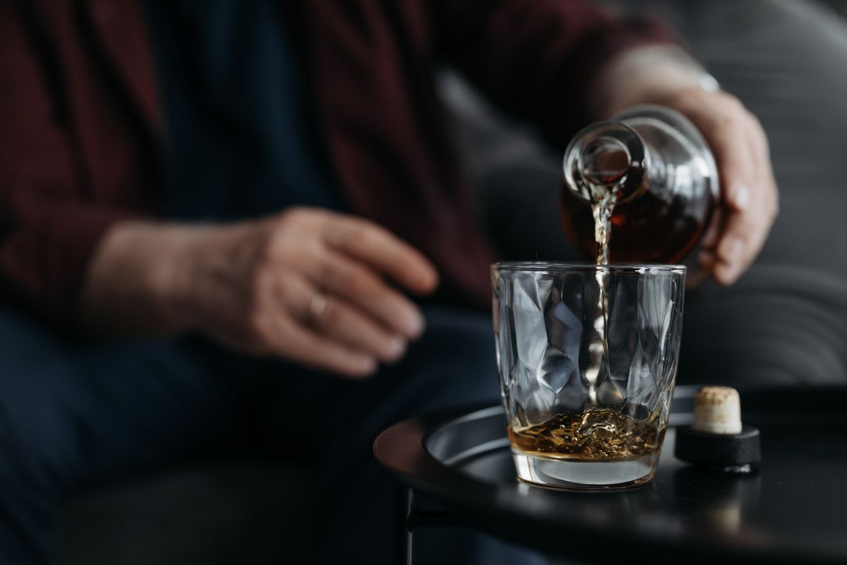 An adult pours whiskey from a bottle into a glass on a table. Indoors setting.