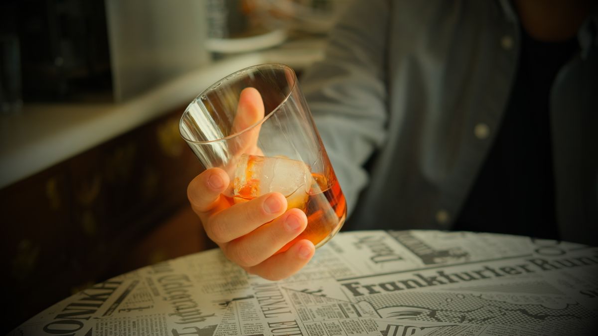 A person holds a whiskey glass on a newspaper-covered table, creating a cozy indoor scene.
