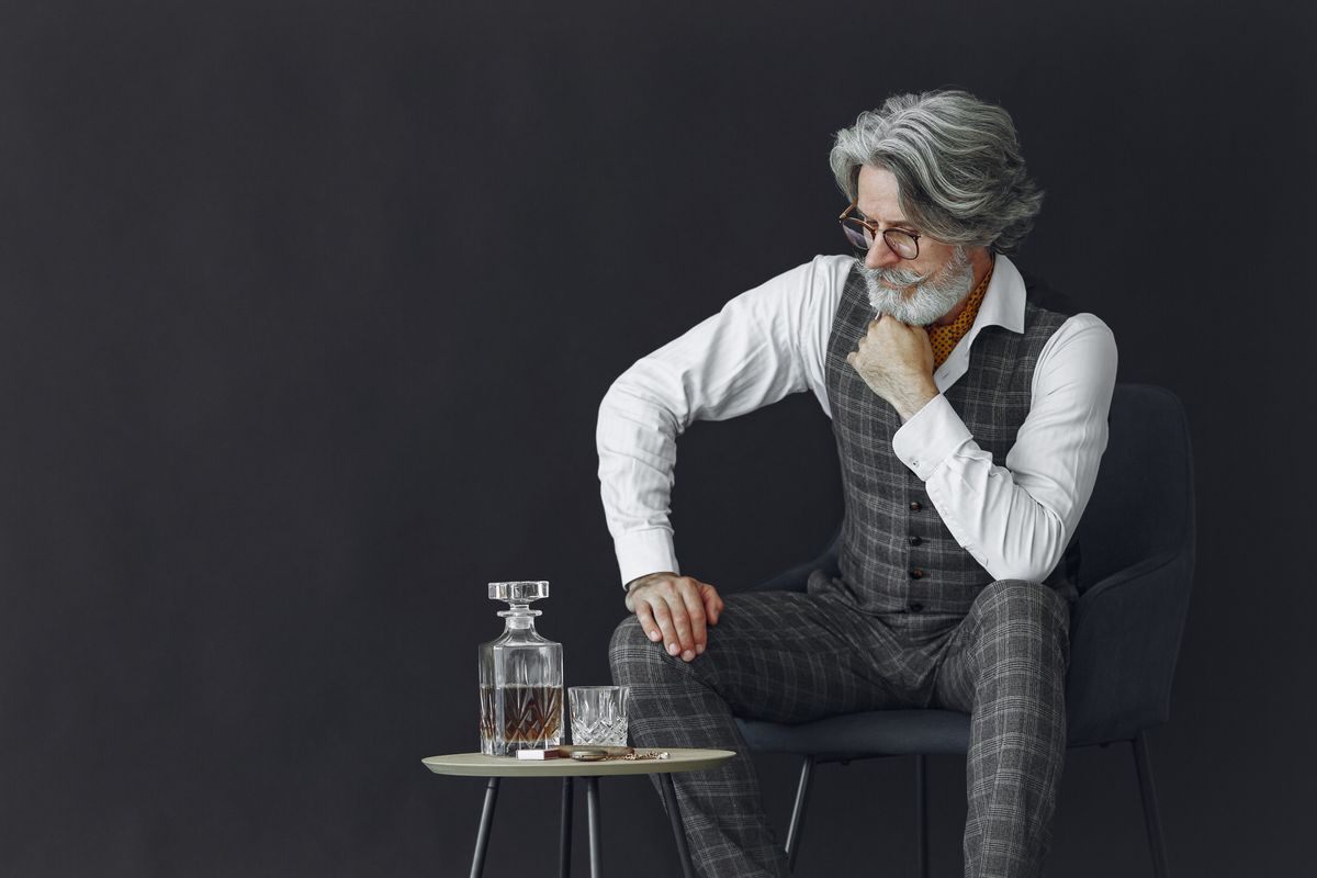 Elegant mature man in a gray suit seated thoughtfully with a whiskey decanter and glass on a table.