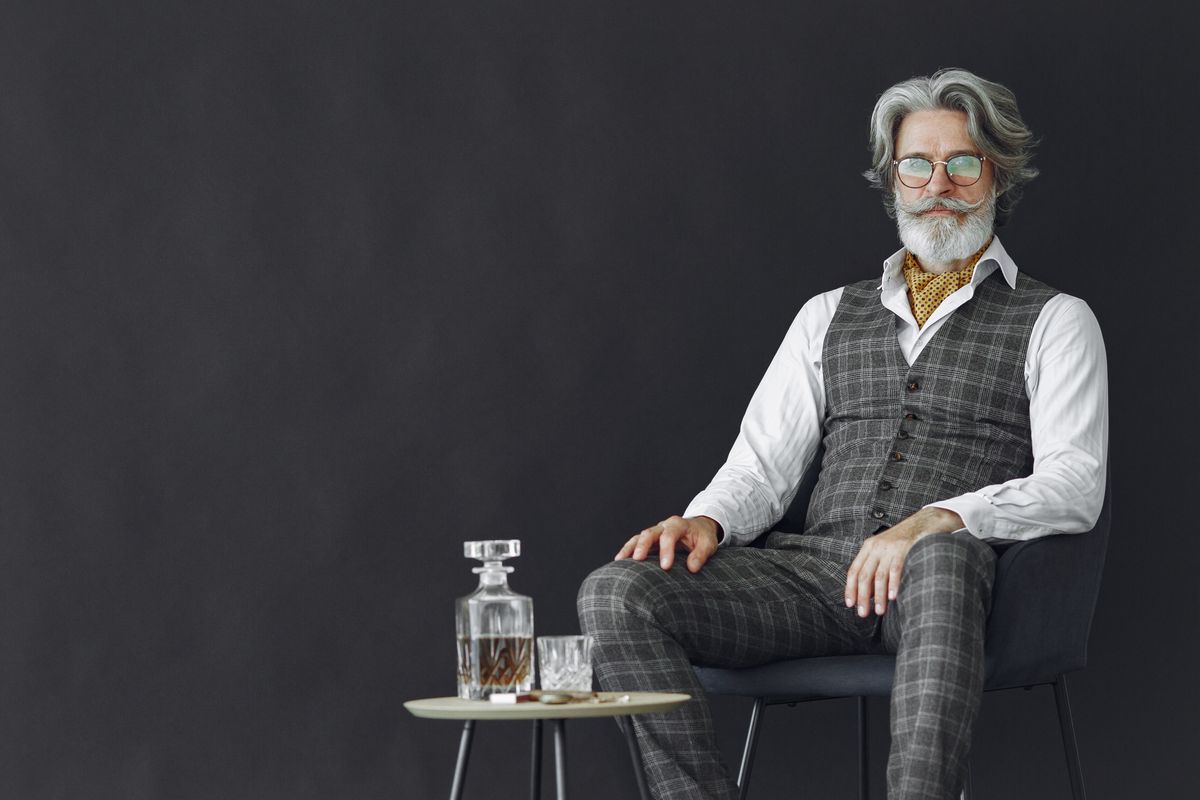 A stylish senior man in a plaid suit sitting with a whisky glass on a table.