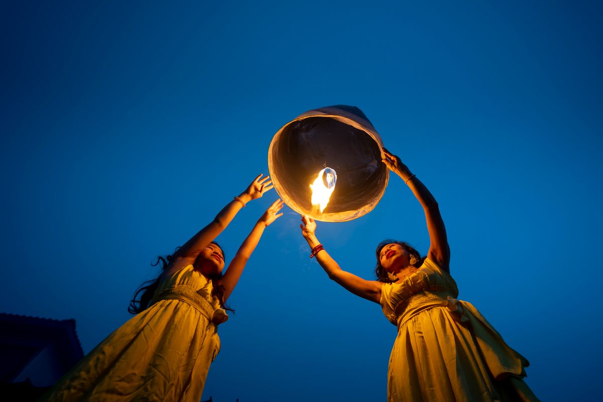 Two women in yellow dresses releasing a glowing sky lantern into the evening sky, symbolizing hope and celebration.