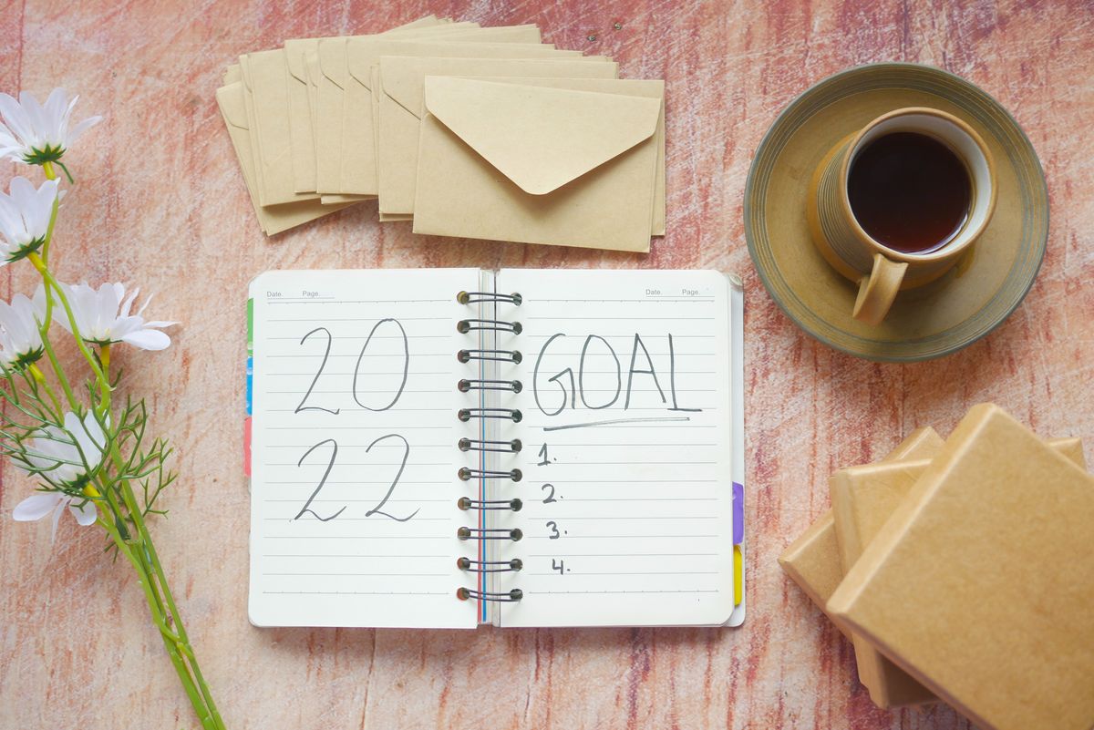 Flat lay of a 2022 goal notebook with coffee, envelopes, and flowers on a rustic desk.