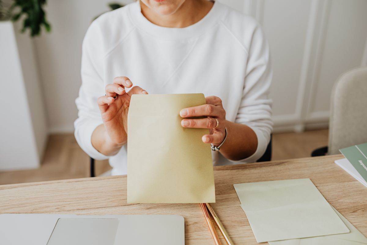 Close-up of a woman sealing a brown envelope while seated at a wooden desk indoors.
