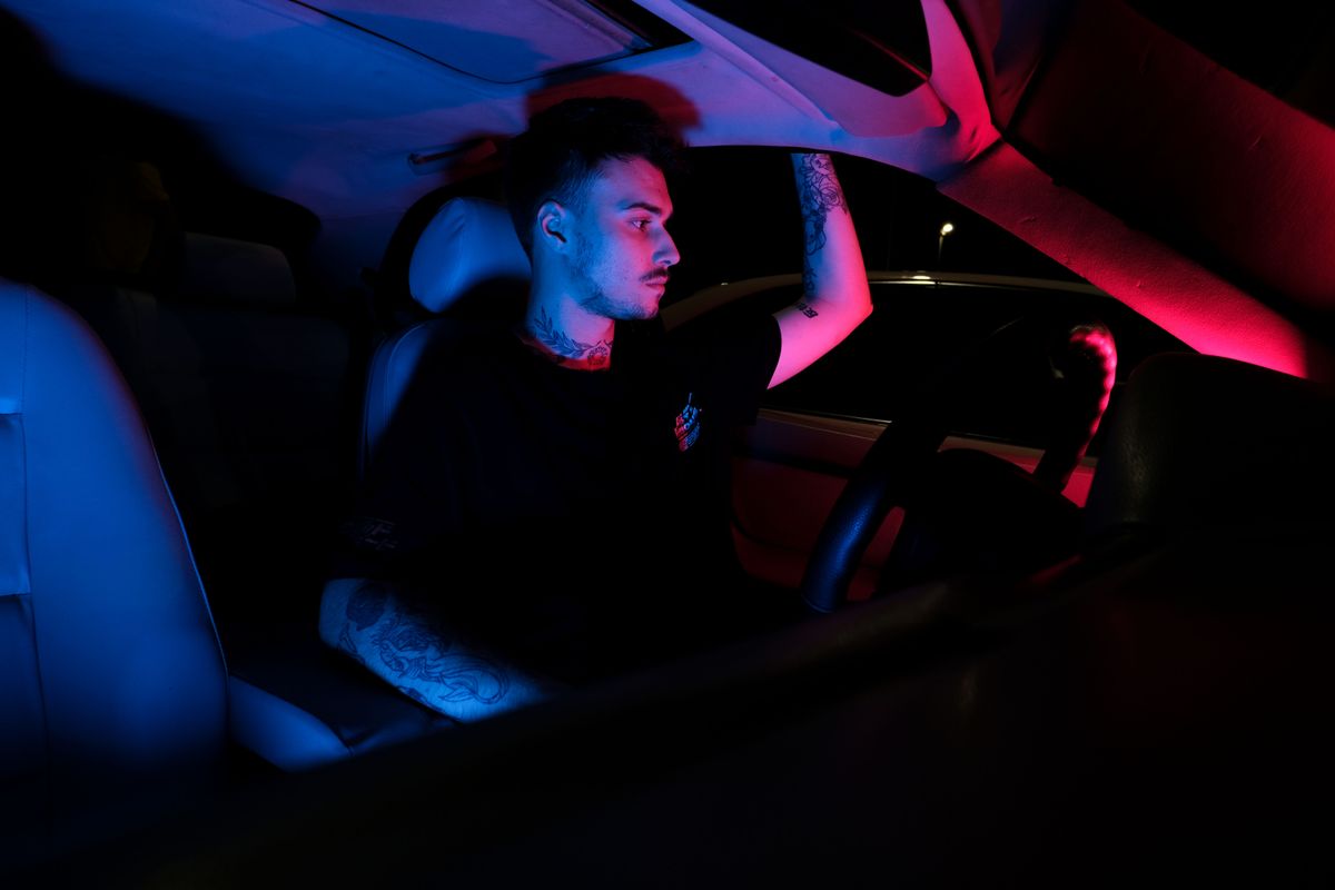 A man with tattoos driving at night, illuminated by artistic red and blue lighting, in a car's front seat.