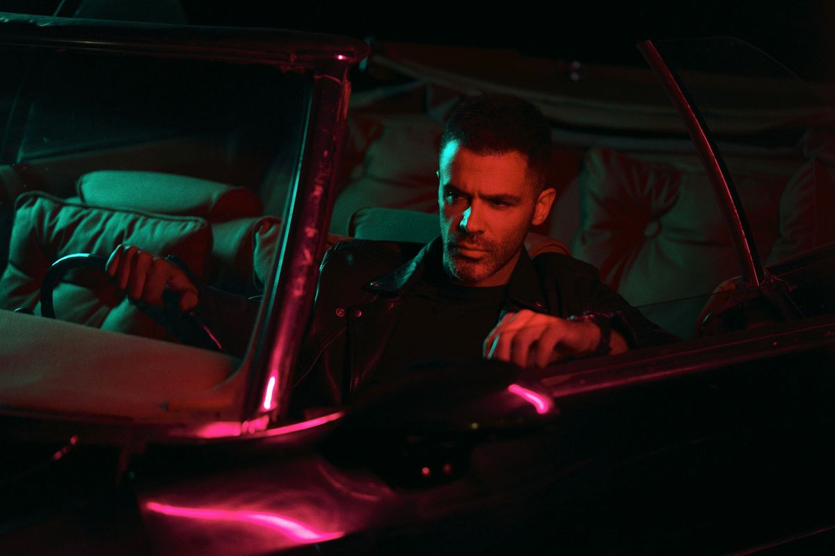 Man in leather jacket driving a vintage car under neon lights.