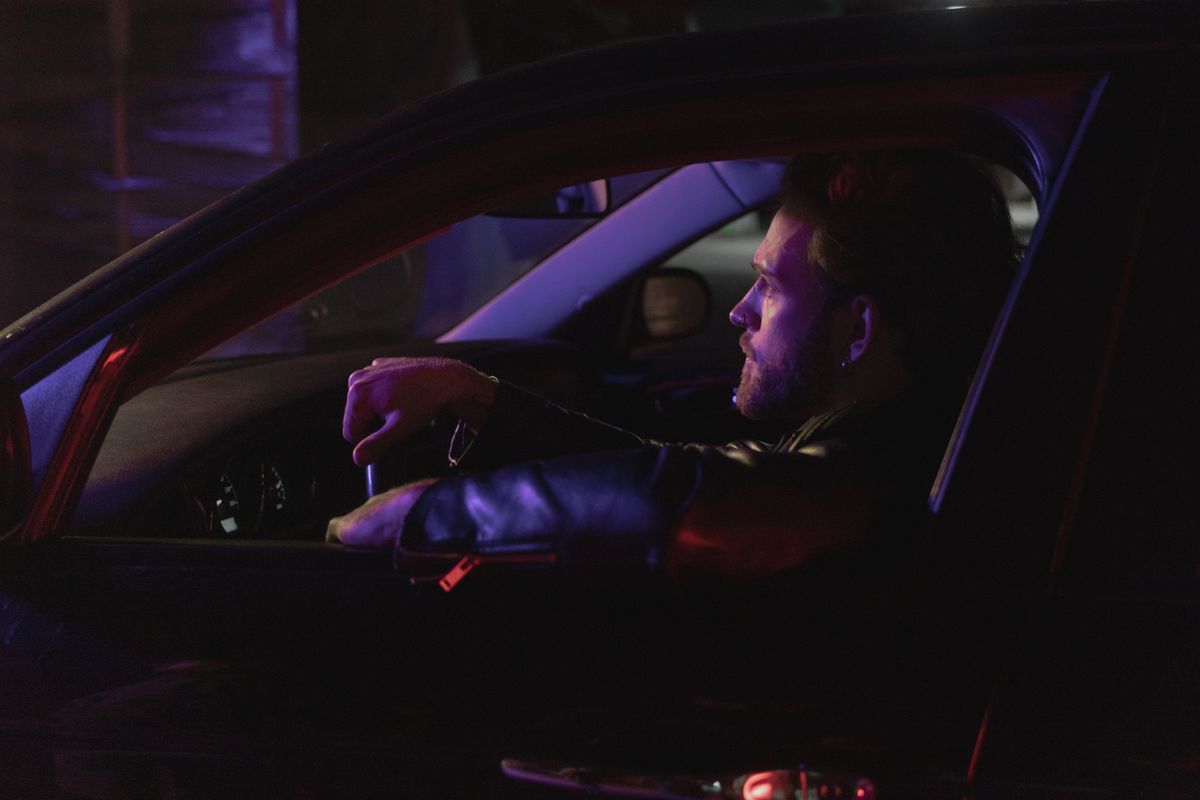 Side profile of a man driving at night in a dark car interior, wearing a leather jacket.