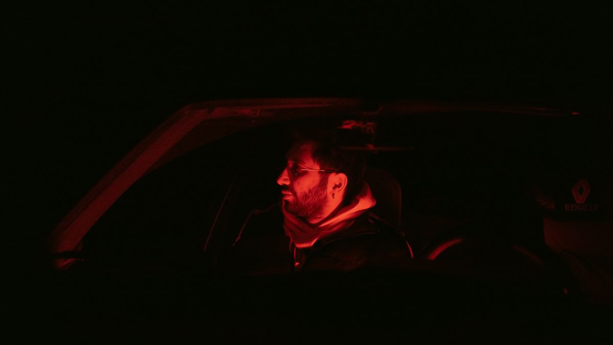 A man sits in a car under red ambient light at night, creating a mysterious atmosphere.