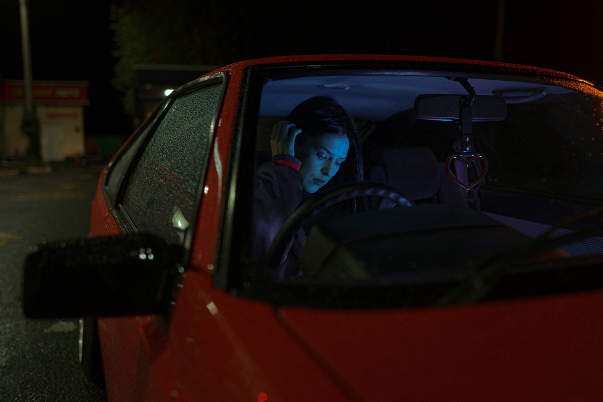 A woman sits alone in a car at night, illuminated by interior lighting, creating a moody and atmospheric scene.