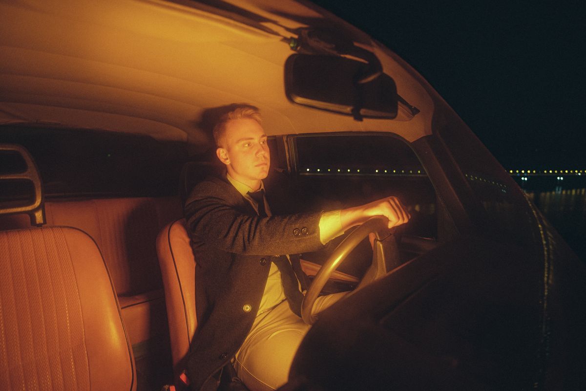 A man in a suit driving a classic car at night under warm lighting.