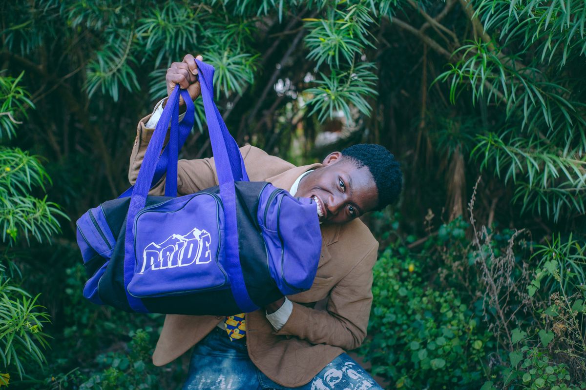 Young man playfully holding a purple duffel bag in a lush, green outdoor setting.