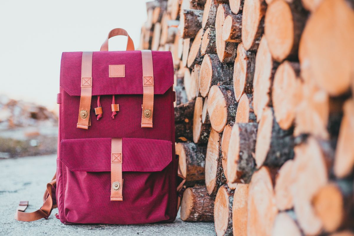 Maroon backpack with leather straps against a rustic log stack backdrop. Ideal for travel and adventure.