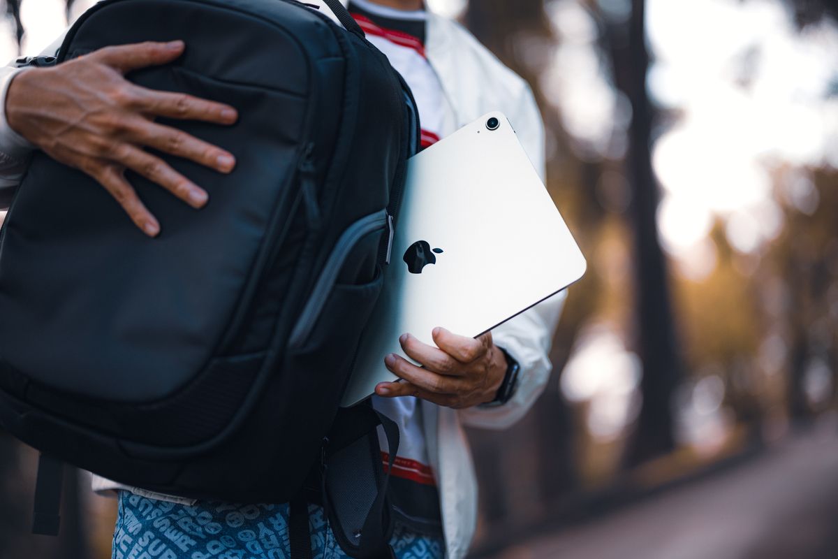 Person holding a tablet near a backpack with a blurred outdoor background, highlighting technology and portability.