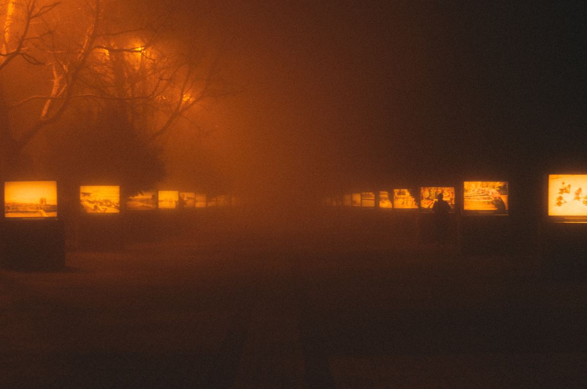 A foggy city boulevard at night with glowing streetlights and outdoor exhibits creating a mysterious ambiance.