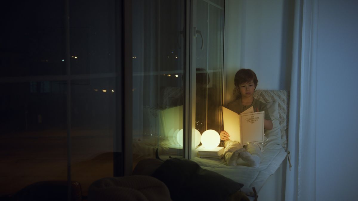 A young boy sits by a window, reading a book with a glowing lamp at night.