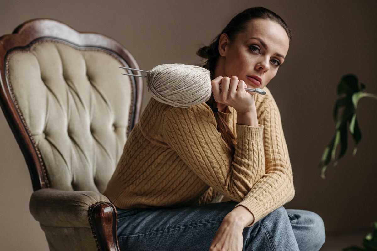 A woman in a cozy knitted sweater with yarn and knitting needles, seated in a vintage chair.