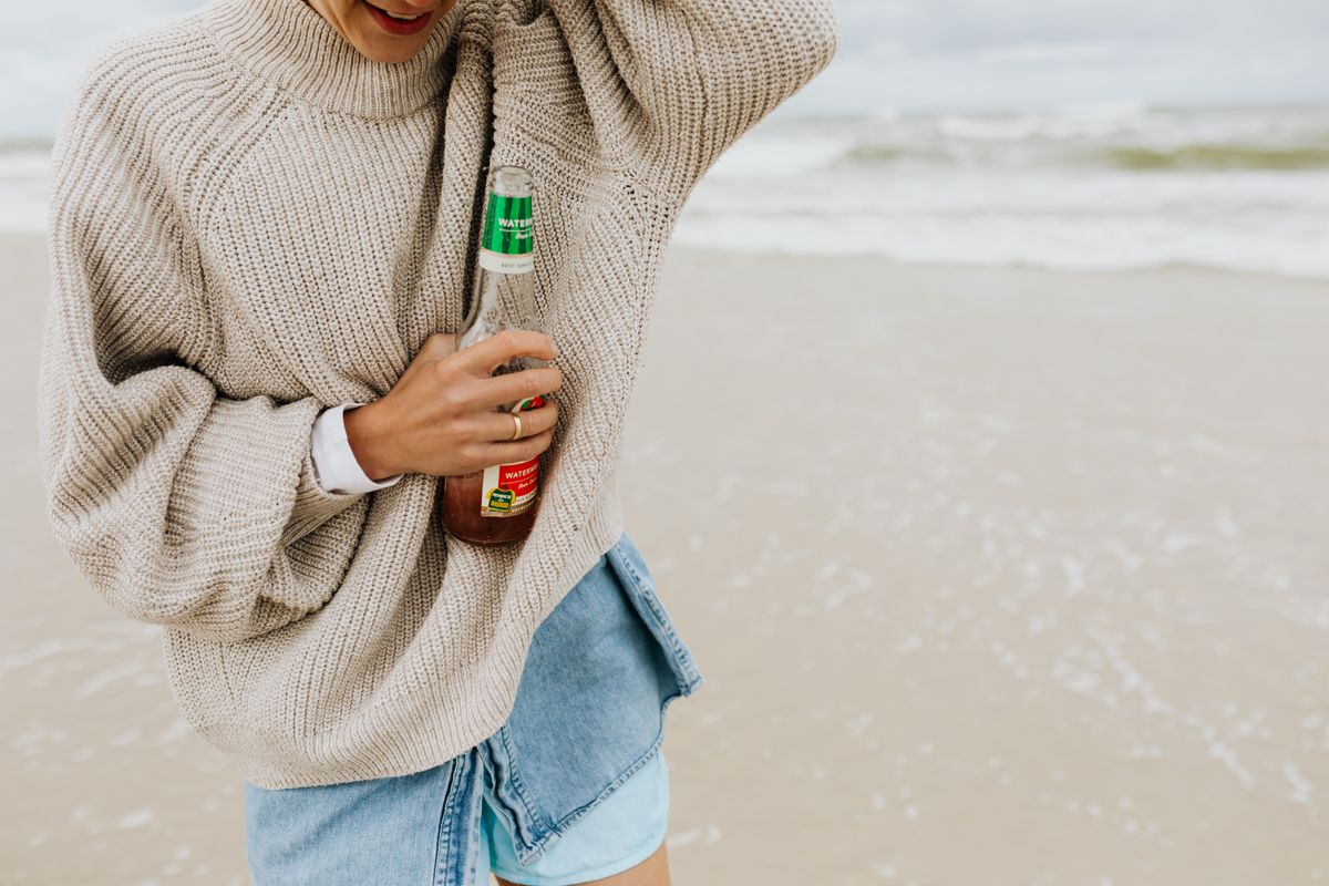 Smiling young woman in a cozy sweater holds a beer bottle at the beach, enjoying a relaxing day.