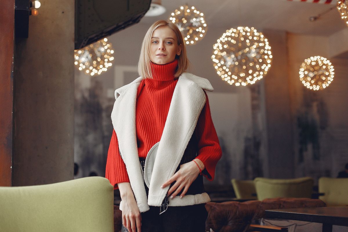 Fashionable woman in red sweater and vest inside a cozy cafe with ambient lighting.
