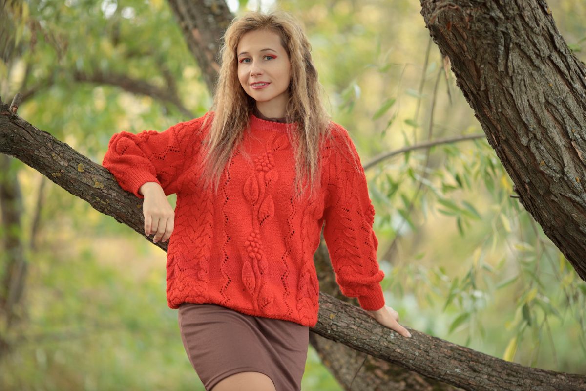 A young woman in a red sweater leaning against a tree trunk in a lush outdoor setting.