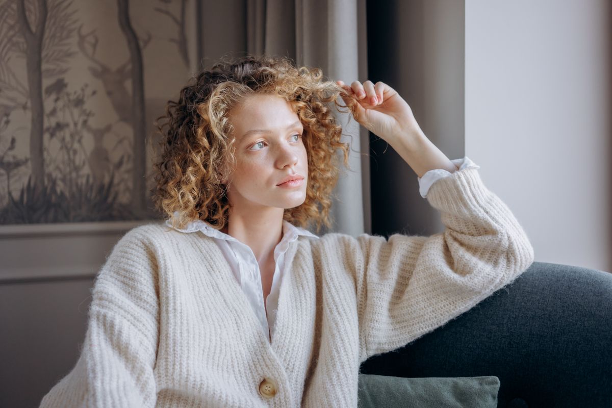 A thoughtful woman in a knitted sweater sitting on a couch in a cozy room.