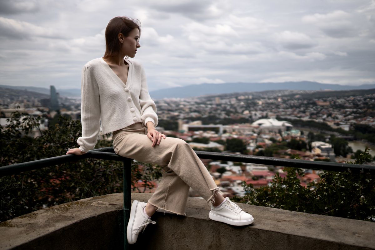 Woman sitting on a rail with city view, wearing casual outfit, serene expression.
