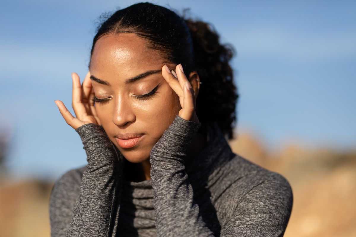 Close-up of a woman in a gray sweater holding her temples, experiencing a headache outdoors in the sunlight.