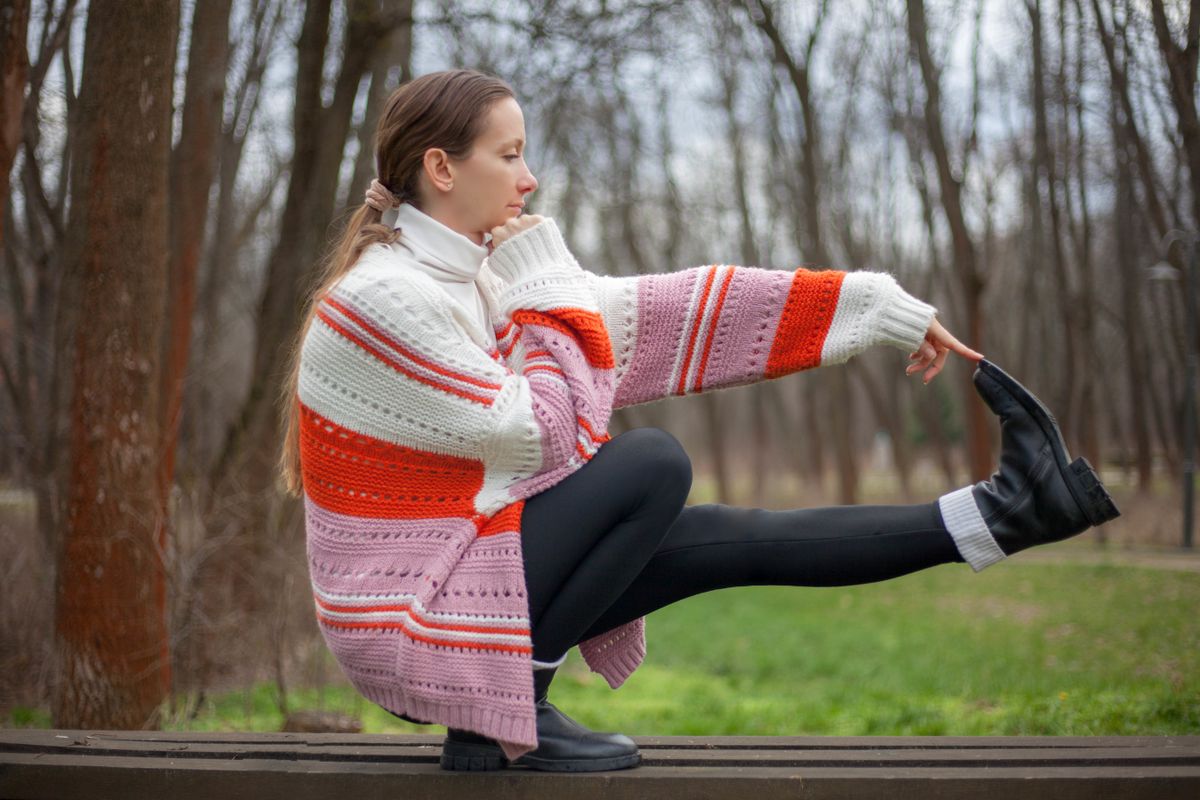 A woman in a colorful sweater practicing a balance pose outdoors in nature.