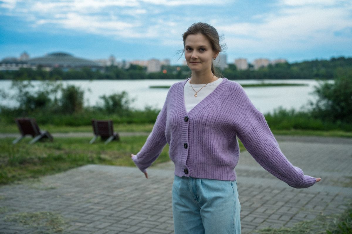 A young woman in a purple sweater poses by a lake with a cityscape backdrop.