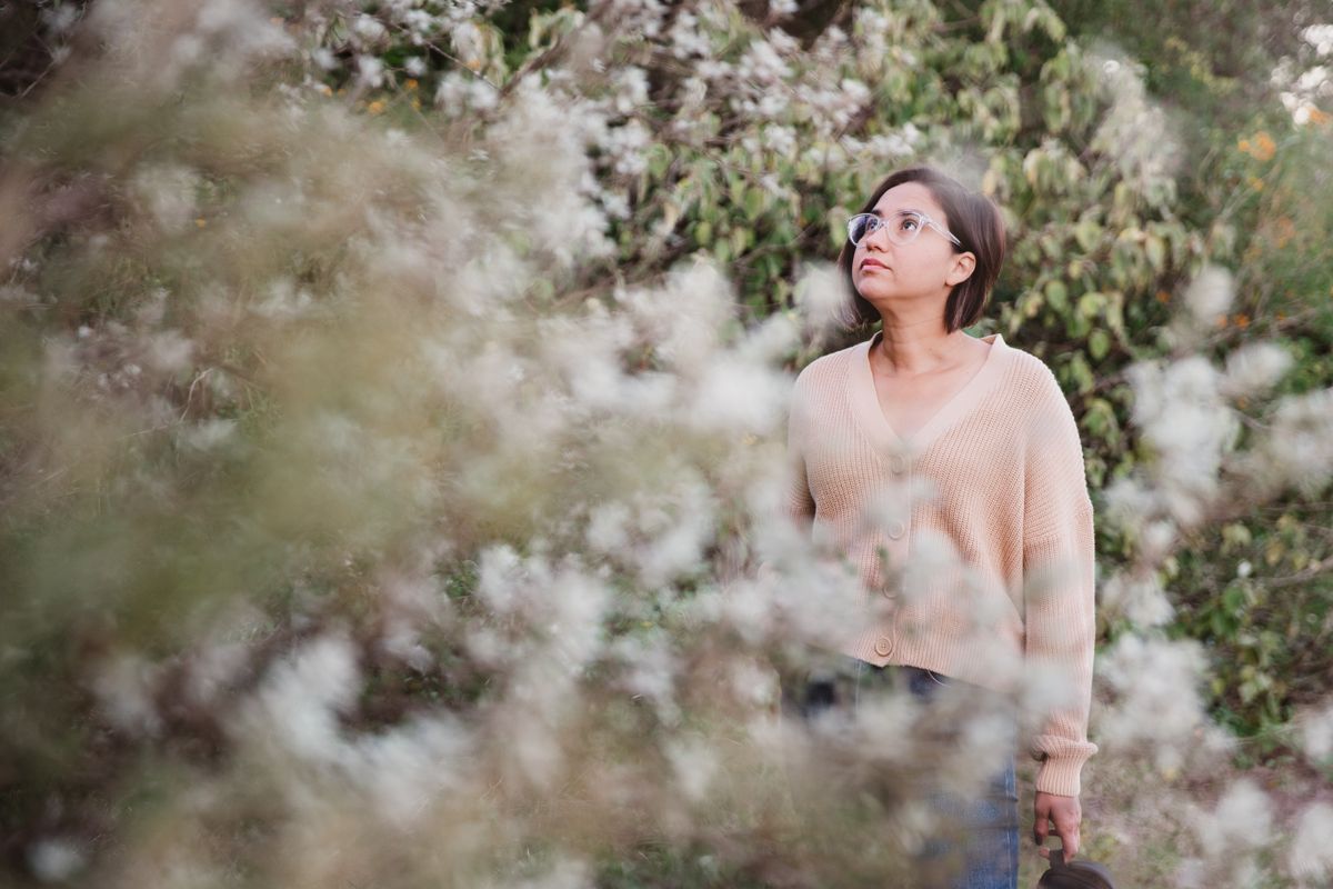 A woman in a cardigan stands amidst blossoming bushes, contemplating nature.
