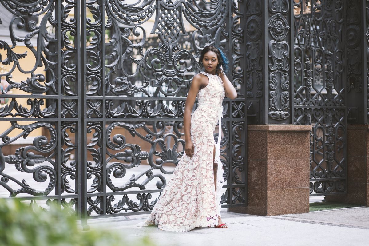 A stylish woman in an elegant lace dress posing against a decorative metal gate outdoors.