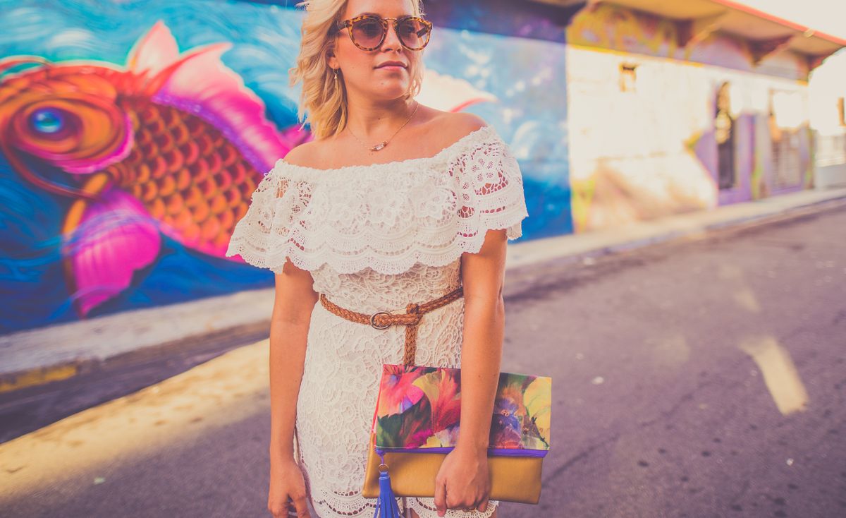 Fashionable woman in a lace dress with a colorful clutch stands near a lively street mural.