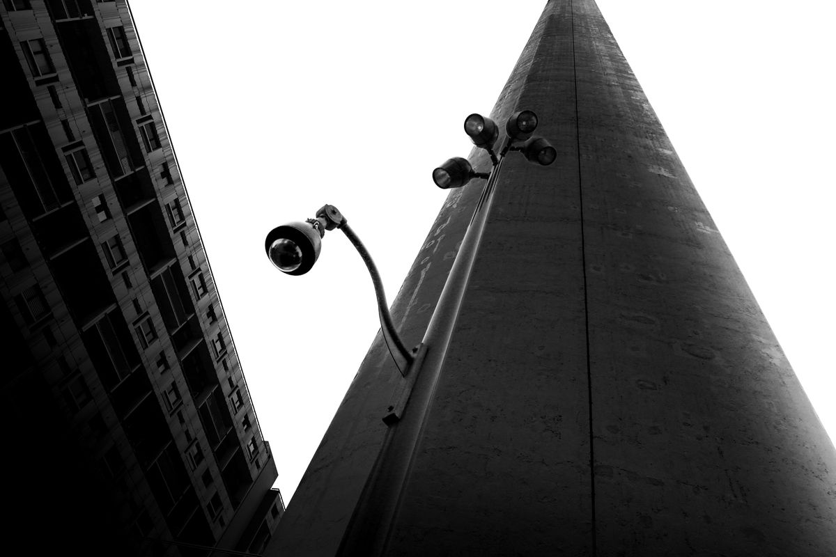 Black and white photo of a streetlight and security camera against a modern building and sky.