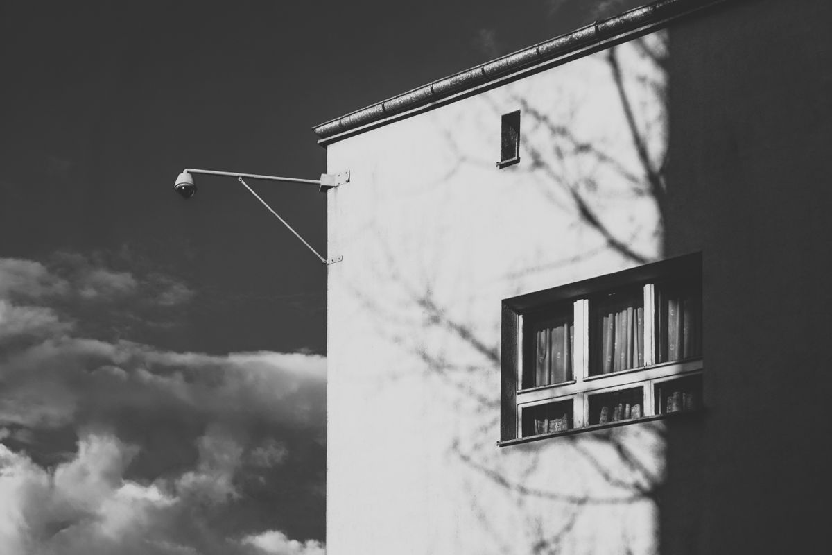 Black and white image of a building with shadows and a security camera against a cloudy sky.