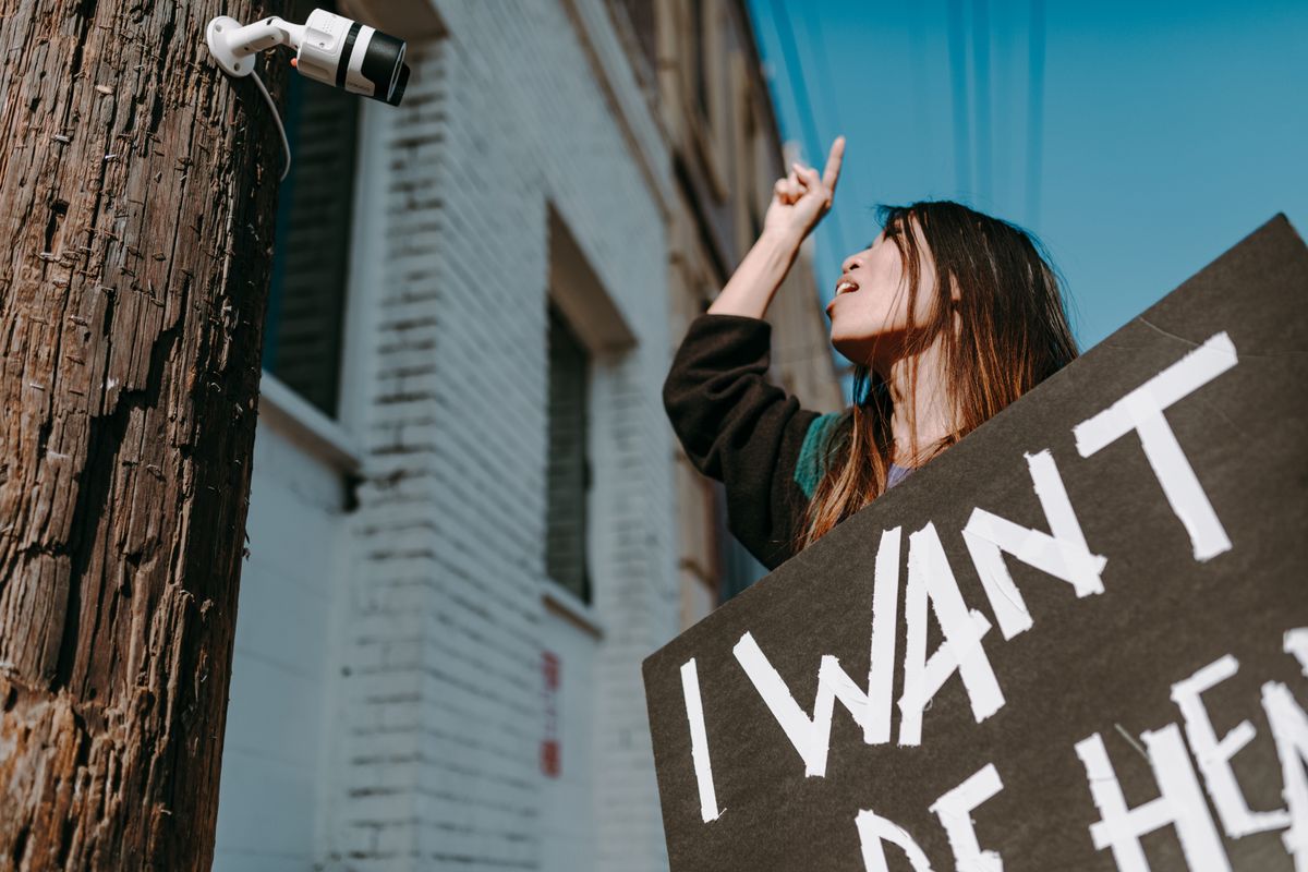 A woman gestures toward a CCTV camera while holding a protest sign in a city setting.