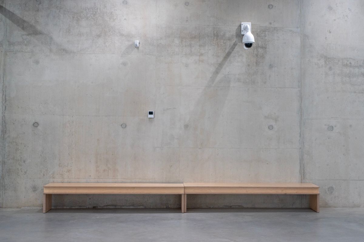 A simple but elegant scene of a wooden bench against a concrete wall with a security camera.