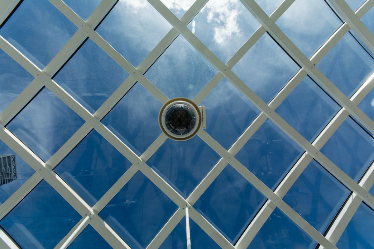 Surveillance camera mounted on a modern glass ceiling with sky reflections.