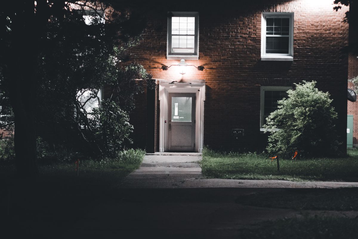 A warmly lit brick house entrance under a night sky, featuring a serene garden setting.