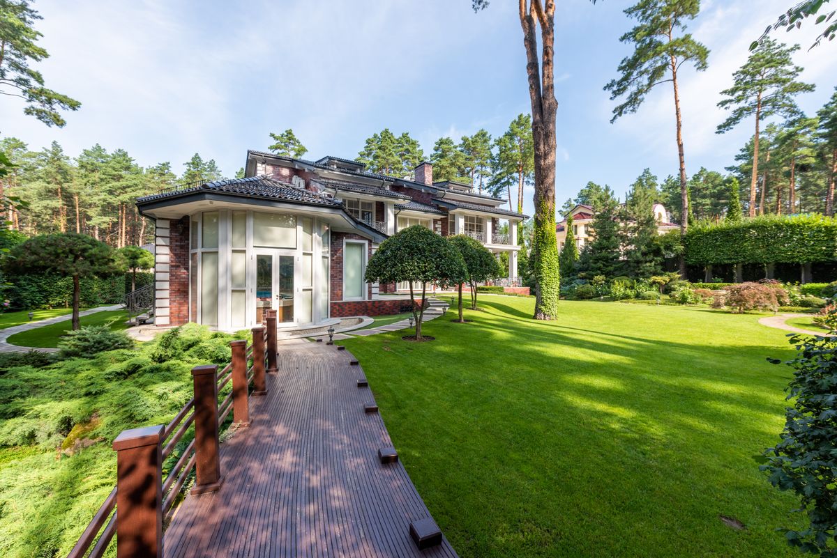Modern house near backyard with wooden path near green grass and plants with trees under blue sky in summer sunny day