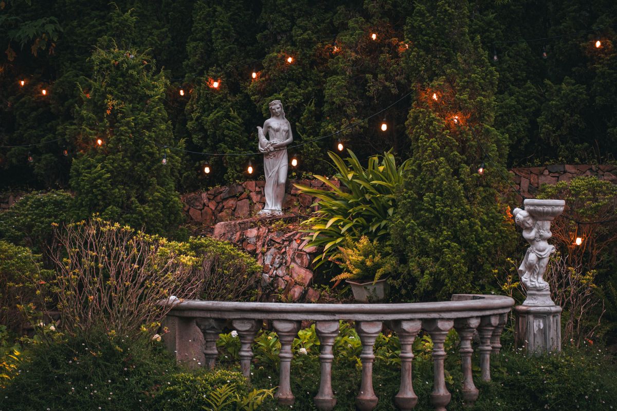 Idyllic garden featuring classical statues and glowing string lights at twilight.