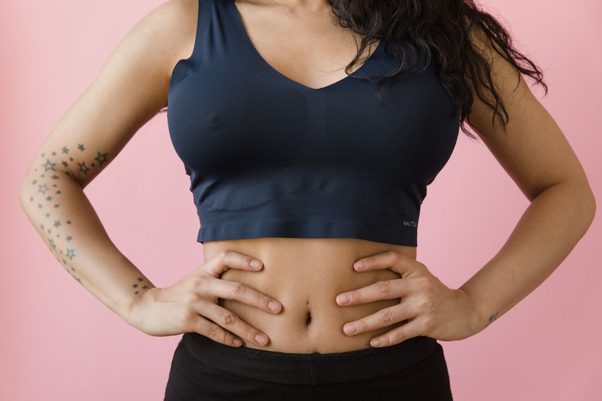 Woman wearing a sports bra and leggings, showing tattoos against a pink background.