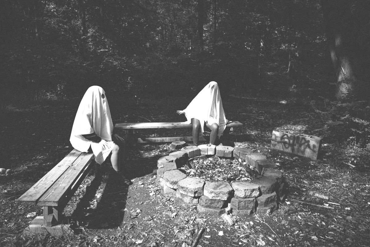 Two people in ghost costumes sit by a fire pit in a forest, creating a spooky Halloween vibe.