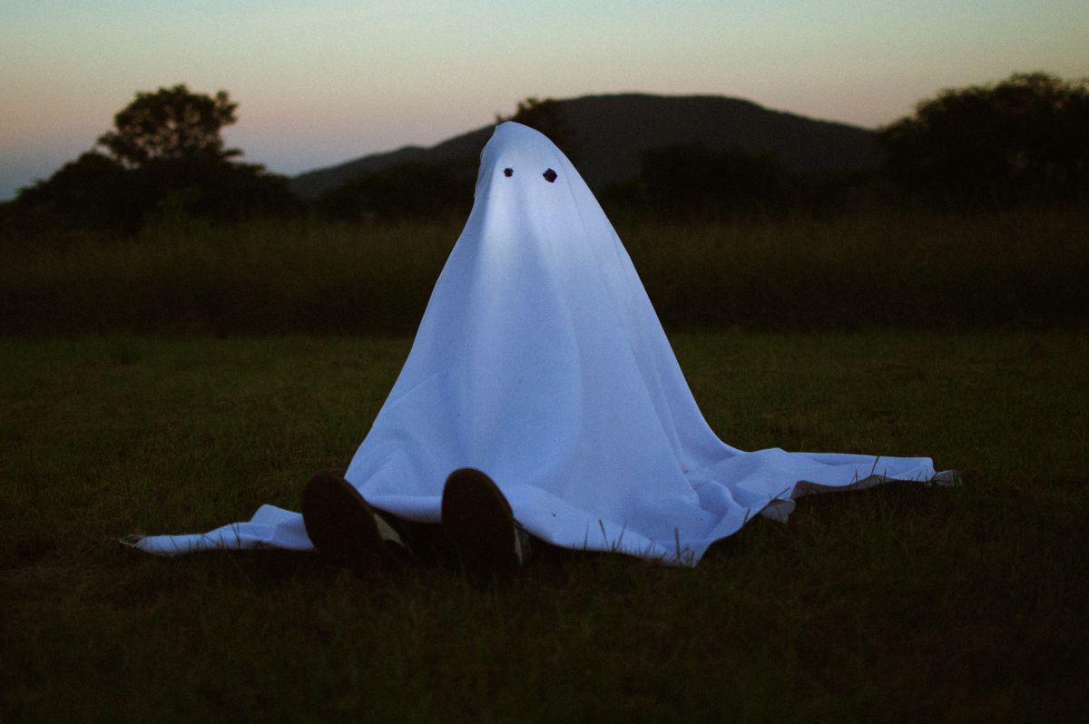A person in a ghost costume sits on a field during dusk, creating an eerie atmosphere.